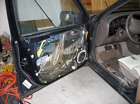 A photo of a car door that's open with the interior panel removed which exposes the speaker and mechanisms inside the door which are mostly covered by a translucent plastic sheet.