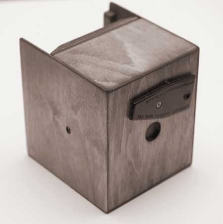 A photograph of a wooden pinhole camera. The camera is a cube that's a little higher than it is wide or deep. The front face of the camera is facing the right edge of the frame. It has a circle in the middle about the size of a dine and a piece of leather that's screwed in above it that acts as a shutter buy rotating it up and down in front of the pinhole which is cut in the middle of the circle. There is also a small circle on the side face of the camera that's screw hole for a tripod mount.