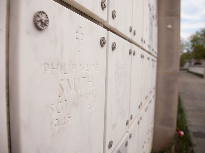 A photo of my dad's nook in a columbarium at Arlington. The camera is right up against the surface angled to look down the length of the structure. The caps are tan marble with names on them that blur as the farther away from the camera they are. Each cap is secured by four ornate rivets in the the corners. Right right quarter of the frame shows the sidewalk leading to the columbarium with a partly cloudy blue sky above it that's casting a soft light on the scene.