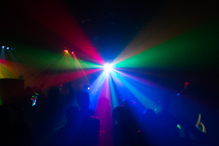An image taken looking towards a multi-colors spot-light with alternating rays of green and red emanating outward to the edges of the frame at random intervals. The silhouette of a crowd takes up the lower portion of the frame with an overall blue haze permeating the scene