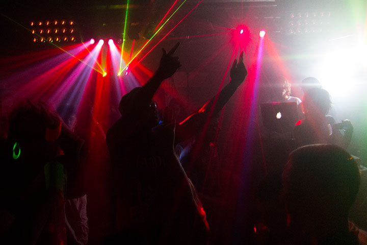 A silhouette of a raver in the middle of a dance floor backed with red spot lights and green lasers. To the right of the image is an area bathed in white light that is backlighting the DJ