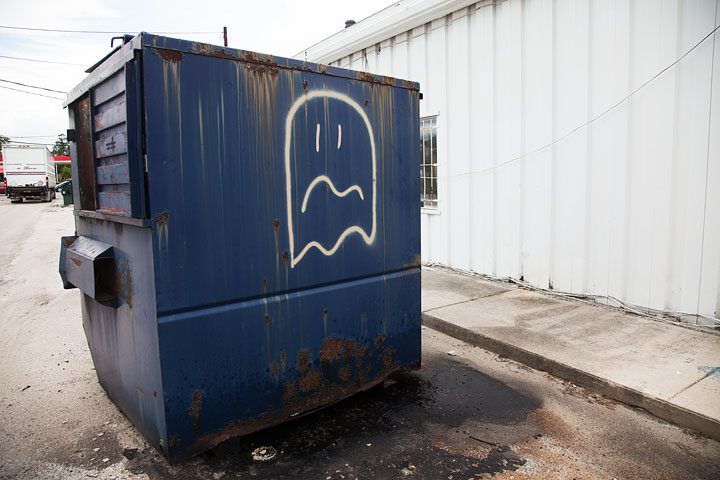 A blue trash dumpster behind a white corrugated steel building with a Pac-Man ghost spray painted on it in white. The ghost's eyes and mouth make it look concerned/distressed