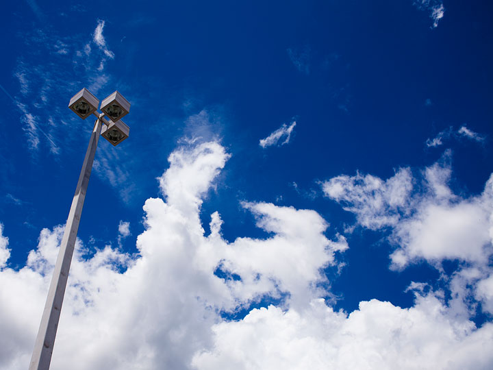 Bright white clouds against a clear, deep blue sky with a lamppost extending up from the lower left portion of the frame. The clouds fill the bottom half of the frame with the sky visible in the upper half. The line between them is a ragged movement up and down from the shape of the clouds. The light post is a square post made of a sun-baked gray metal. Three square boxes extend from the top of the post at ninety degree angles. Each contains a a circular light.