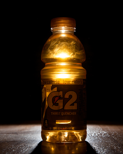 A photo of a 20 ounce orange Gatorade G2 bottle. The only light in the scene is directly behind the bottle which results in a glow from the center of the liquid and refleced highlights expanding out across the dark wooden table it's sitting on. The background is completely black staring just above the bottom part of the label.