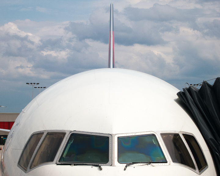 A plane parked at an airport gate. The image is taken from directly in front of the plane with only the windows and above visible and the body of the plane forming a half circle. A black canvas looking attachment that connects the plane to the gateway rests on the plane on the right side of the image. The tail fin is sticking straight up at the back with a partly cloudy sky behind it.