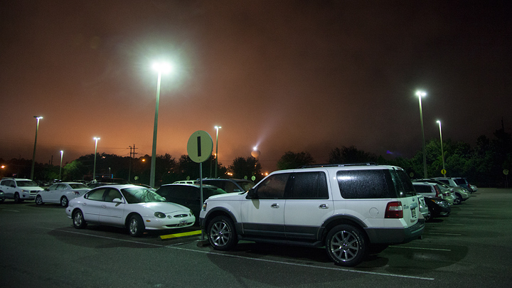Cars in an airport parking lot under an overcast sky before dawn. The cars are illuminated by the parking lot lights with the orange sodium vapor lights of the airport area behind them coloring the sky.