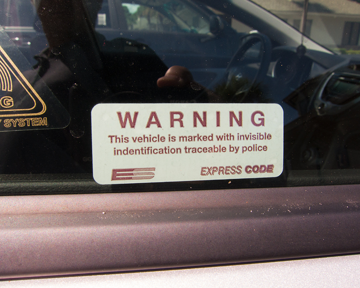 A sticker on the side window of a car that reads 'WARNING This vehicle is marked with invisible identification tracable by police' with the name 'Express Code' under it