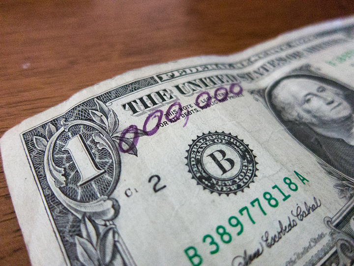 The upper left corner of a dollar bill laying on a wodden table. Someone has drawn six zeros after the 1 in the corner to turn the number into 1 million.