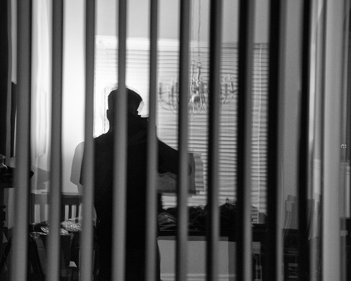 A self-portrait taking as a backlit reflection in a window with open vertical blinds on it. The blinds make dark lines extending up and down the image which is lit by a soft light opposite wall. The image is cropped square. Three quarters of my silhouette extends from the bottom of the frame up until just below the top third of the image where I'm move one body position off center to the left. My elbow is extended to the middle of the frame as I hold the camera. 